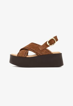 Sandalia plataforma de ante marrón con tiras cruzadas, detalles perforados, hebilla dorada ajustable y suela gruesa negra.