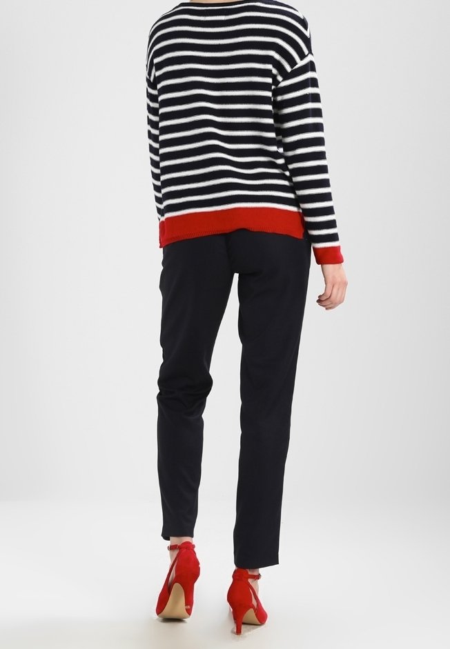 Kobieta ubrana w czarne spodnie, czarno-biały sweter w paski z czerwonymi mankietami oraz czerwone buty na wysokim obcasie, stojąca tyłem.