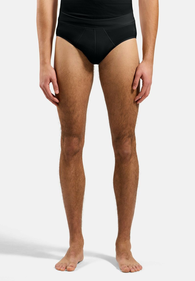 Mann, der barfuß steht und schwarze, hoch taillierte Slip und ein schwarzes Oberteil trägt, vor einem schlichten hellen Hintergrund.