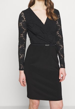 Vrouw die een zwarte jurk tot op de knie draagt met lange mouwen van kant en een overslagtop, aangesnoerd in de taille met een dunne tailleband.