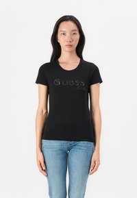 Black short-sleeve T-shirt with a crew neckline and glittered "Guess est. 1981" logo on the front, paired with blue jeans.