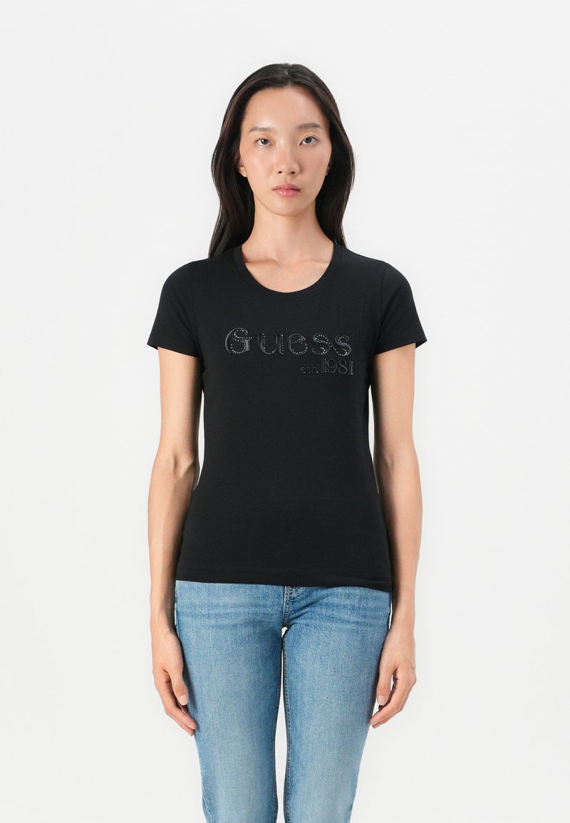Black short-sleeve T-shirt with a crew neckline and glittered "Guess est. 1981" logo on the front, paired with blue jeans.