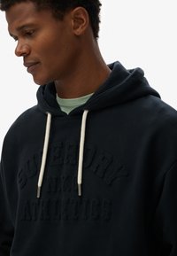 Sudadera con capucha de color azul marino, hecha de un tejido suave, con un logo en relieve de un azul más oscuro, cordones blancos y una camiseta interior de color verde menta.