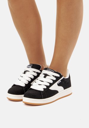 Zapatillas de ante negras con cordones blancos y un acento en blanco. Suela de goma plana en color marrón claro. Presentadas de frente, mostrando detalles y construcción.