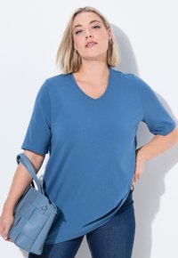T-shirt blu con scollo a V e maniche corte, realizzato in morbido materiale, abbinato a una borsa intrecciata di colore azzurro chiaro, su sfondo bianco.