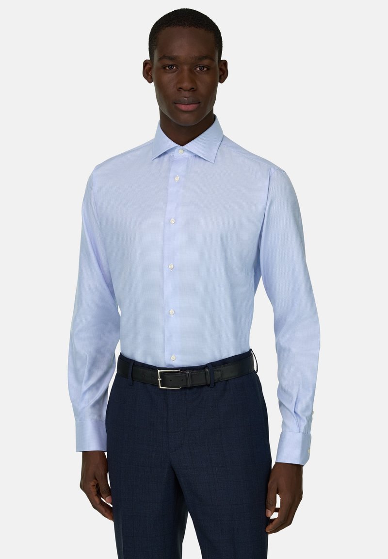 Homme portant une chemise habillée bleu clair à boutons, rentrée dans un pantalon bleu marine foncé avec une ceinture noire, debout face à l'avant sur un fond blanc.