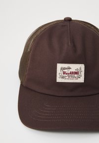 Barna sapka, szilárd anyagból készült elülső résszel és hálós hátsó résszel. Téglalap alakú felvarróval rendelkezik, amelyen a "BILLABONG" felirat és díszítő elemek találhatók. Lapos karima.