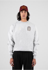 Felpa grigia con design ricamato, che presenta uno stemma e il numero 3. Abbinata a pantaloni neri plissettati e a un cappellino bordeaux.