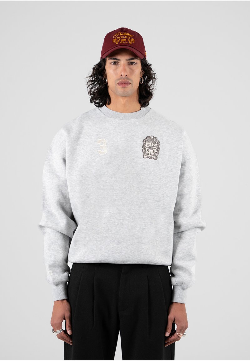Felpa grigia con design ricamato, che presenta uno stemma e il numero 3. Abbinata a pantaloni neri plissettati e a un cappellino bordeaux.