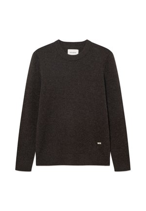 Jeune homme debout, mains derrière le dos, portant un pull marron, un jean bleu, des chaussures noires et une casquette beige sur fond blanc.