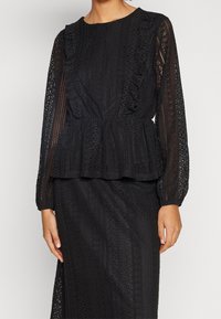 Top in pizzo nero con maniche lunghe e accenti arricciati, modelli testurizzati e vita peplum. Presenta un collo rotondo e dettagli intricati.