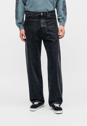 Person trägt lockere schwarze Jeans mit Muster, schwarz-weiße Sneakers und ein hellblaues Langarmhemd mit dunklen Designs auf den Ärmeln.