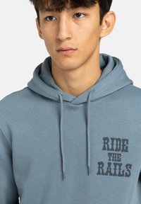 Felpa leggera azzurra realizzata in tessuto morbido, con cappuccio regolabile e una stampa grafica del testo "RIDE THE RAILS" in caratteri scuri sul petto.