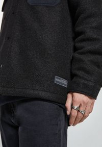 Giacca in misto lana nera con finitura texturizzata, dotata di una tasca sul petto in blu navy e un'etichetta sull'orlo. Sono visibili jeans scuri e anelli.
