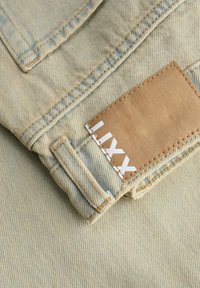 Beżowa tkanina dżinsowa o teksturowanej powierzchni, z brązową skórzaną metką przyszytą w pasie, z białym wytłoczonym napisem "LIXY".
