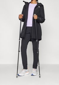 Veste imperméable noire avec fermeture éclair, chemise violette, pantalon noir, baskets grises avec des accents orange et bleu, et bâtons de trekking.