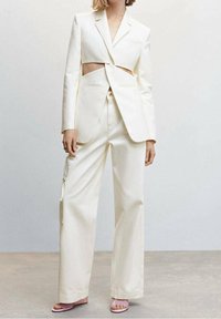 Femme portant un tailleur écru avec des découpes à la taille, un pantalon à jambes larges avec une poche latérale, et des talons ouverts rose.