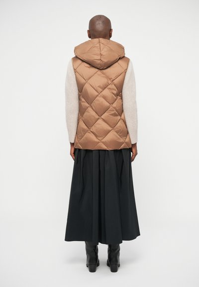 Gilet rembourré matelassé marron avec capuche, porté sur un pull gris clair, associé à une longue jupe noire et des bottes noires jusqu'aux genoux.