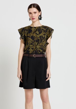 Donna con top fantasia oliva e nero con maniche arricciate, pantaloncini neri con cintura, in piedi davanti a uno sfondo chiaro uniforme.