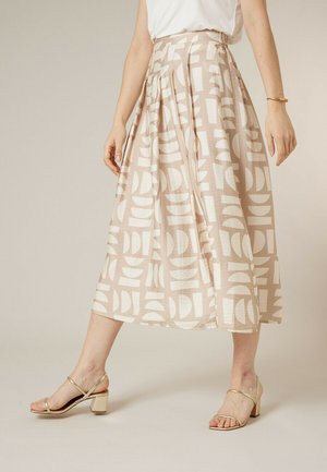 Femme portant une jupe midi beige à motifs géométriques blancs avec des plis, associée à des sandales à talons blocs dorés à bout ouvert et un haut blanc sans manches.