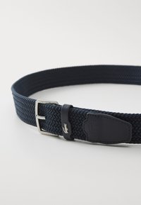Navy woven belt made of elastic material, features a silver metal buckle, leather accents, and a small logo emblem.