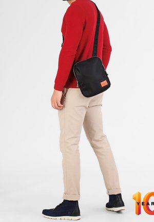 Persoon draagt beige broek, marineblauwe hoge sneakers en een rode trui, en draagt een zwarte crossbody tas met een oranje patch.