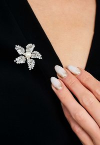 Silver-tone floral brooch with leaf-like details, central pearl, attached to a black garment. Fingernails feature gradient with gold accents.