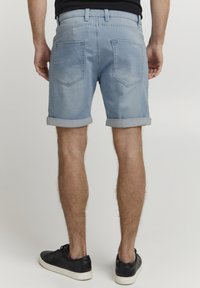 Ljusblå denimshorts med uppvikta fållar, två bakfickor med sömdetaljer, bärs med svarta sneakers, visad från bakre sidan.