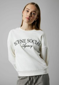 Sudadera blanca con el bordado "SCENE SOCIAL Agency" en negro en script. Presenta un ajuste relajado y puños acanalados. Textura suave.