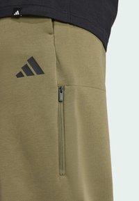 Olijfgroene sportbroek met ritszak, voorzien van een zwart Adidas-logo en een gladde textuur. Ontworpen voor comfort en flexibiliteit.