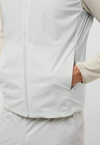 Gilet leggero grigio chiaro, con una texture liscia e una zip frontale. Presenta tasche laterali e una morbida maglietta a maniche lunghe di colore chiaro sotto.