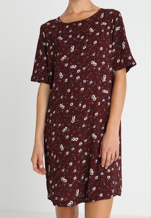 Nainen, jolla on lyhythihainen, polvipituinen burgundinpunainen mekko, jossa on pieni valkoisen ja sinisen kukkakuviointi yksivärisellä taustalla.