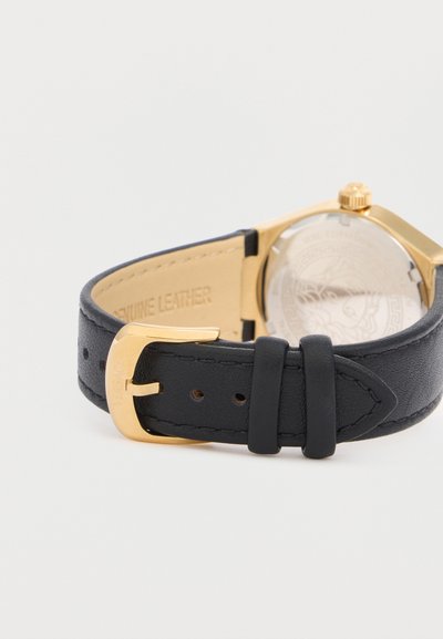 Μαύρο δερμάτινο λουράκι ρολογιού με χρυσή αγκράφα προσαρτημένο σε χρυσή θήκη ρολογιού που δείχνει το χαραγμένο σχέδιο στο πίσω μέρος.