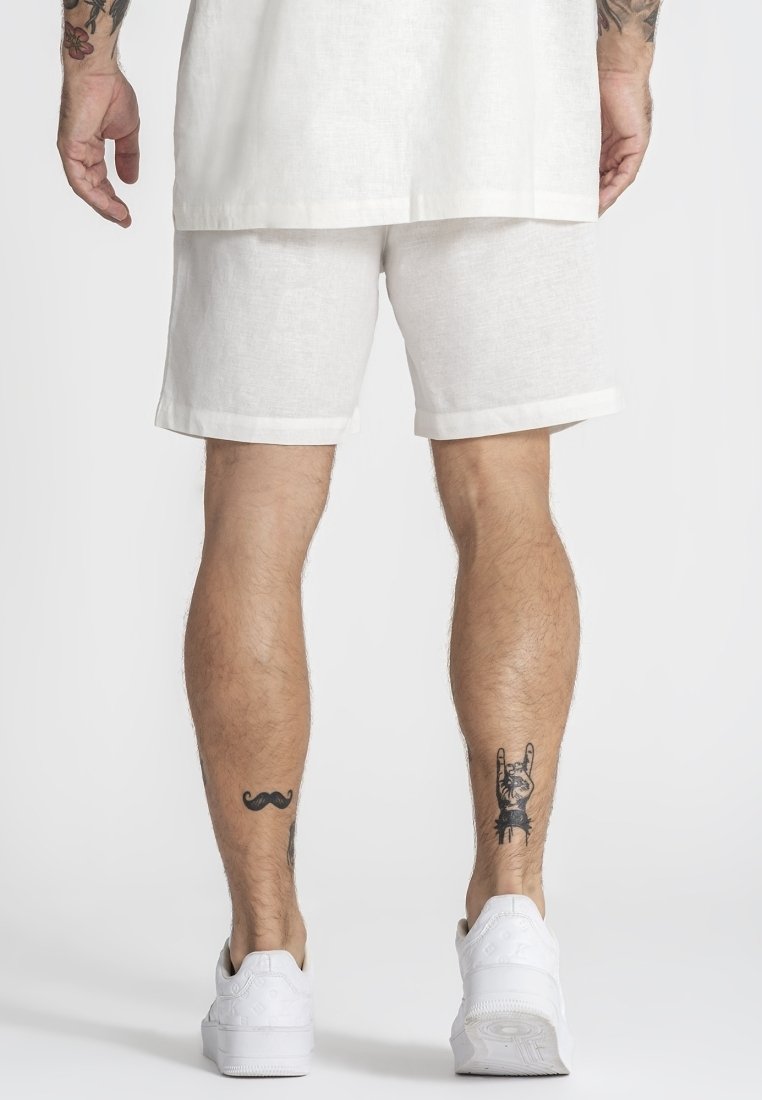Uomo che indossa pantaloncini bianchi e scarpe da ginnastica bianche, con tatuaggi di un baffo e una mano che fa il segno del rock sul retro delle gambe.