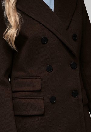 Manteau marron foncé à double boutonnage avec des boutons noirs et deux poches à rabat, porté sur un haut bleu clair, avec de longs cheveux blonds visibles.
