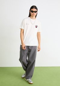 Weißes Baumwoll-T-Shirt mit dem "Champion"-Logo, kombiniert mit grauen weiten Hosen und silbernen Sneakern. Accessoires umfassen schwarze Sonnenbrillen und eine silberne Kette.