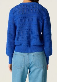 Blå stickad tröja med texturerad mönstring och ribbade muddar. I kombination med ljusblå jeans som visar baksidan av båda plaggen.