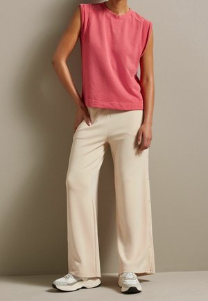 Personne portant un haut rose sans manches, un pantalon large beige avec des boutons sur le côté et des baskets blanches, debout contre un mur beige.