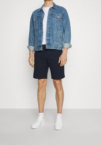Jeansjacka i ljusblått, vit t-shirt med rund halsringning, mörkblå shorts och vita sneakers. Jackan har knappframsida och bröstfickor.