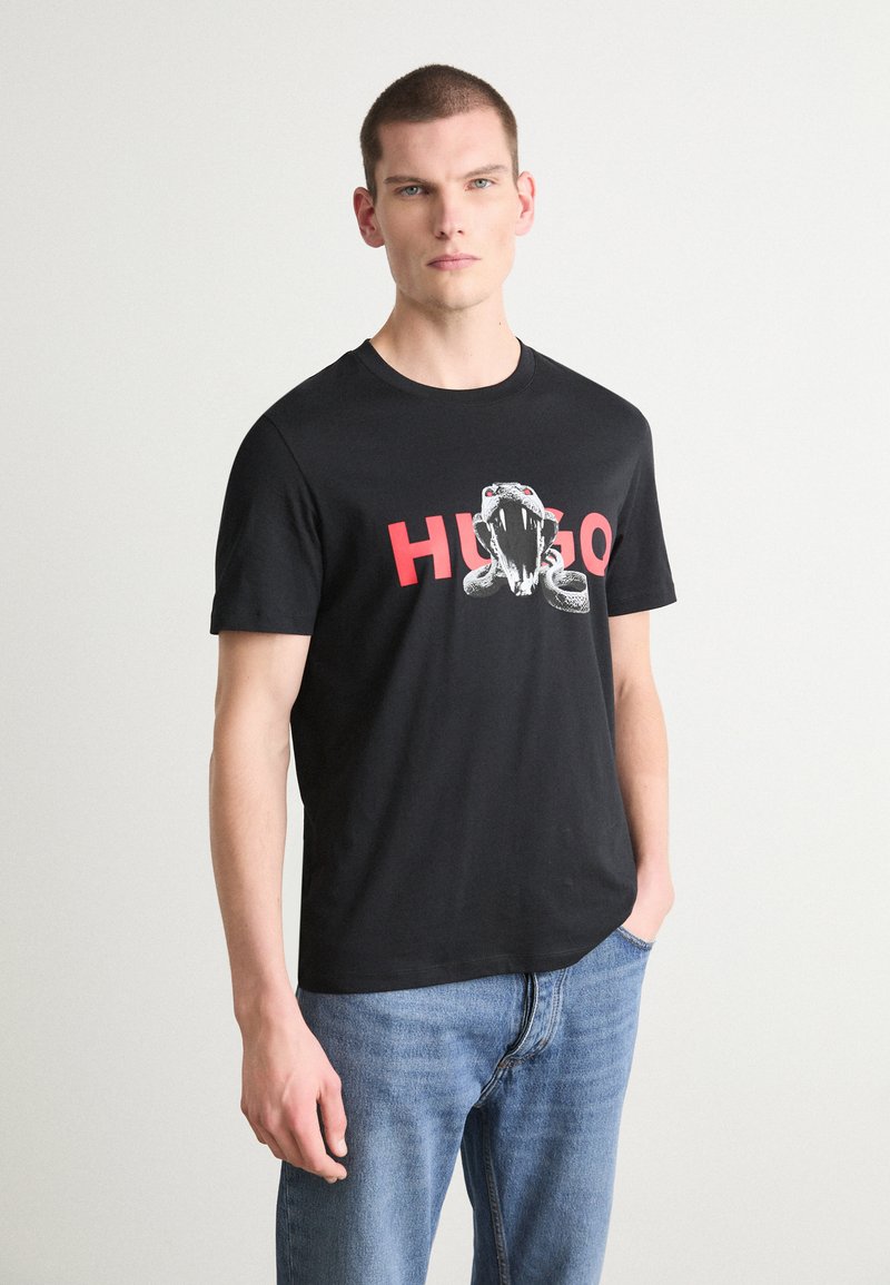 Svart bomull t-skjorte med grafikk av en slange og ordet "HUGO" i rødt. Korte ermer og rund hals, sammen med blå jeans.