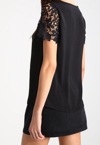 Černý top s krátkými krajkovými rukávy, hladká tkanina, volný střih a zaoblený zadní lem. Kombinován s černou džínovou sukní.