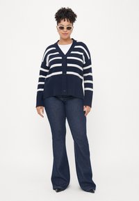 Μπλε ριγωτό καβιού με λευκές ρίγες, κουμπωτό μπροστινό μέρος, V-λαιμόκοψη και ριχτό στρίφωμα, συνδυασμένο με σκοτεινές μπλε φούστες τζιν. Τα αξεσουάρ περιλαμβάνουν γυαλιά ηλίου.