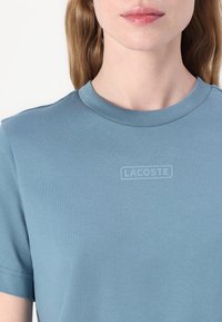 Šviesiai mėlynas medvilninis marškinėlis trumpomis rankovėmis, su ribotu apvaliu kaklu ir embosuotu "LACOSTE" logotipu krūtinėje. Lygus paviršius ir prigludusi forma.