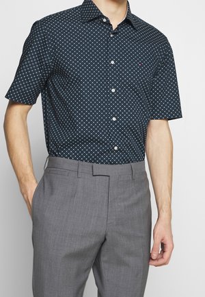 Mann trägt ein marineblaues kurzärmeliges Hemd mit weißen kleinen Kreiseln und graue Anzughose, Hand in der Hosentasche.