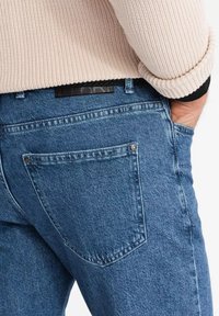 Persona que lleva puesto un pantalón de mezclilla azul y un suéter de punto beige con la mano en el bolsillo, mostrado desde la cintura hasta la mitad del muslo.