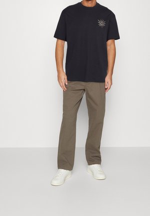 Schwarzes T-Shirt mit kurzen Ärmeln und einer Sonnengrafik auf der Brust, kombiniert mit olivgrünen, geraden Hosen und weißen Sneakers.