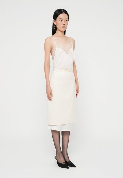 Caraco blanc avec bordure en dentelle, associé à une jupe crayon couleur crème. Talons noirs à bouts pointus et collants noirs transparents complètent le look.