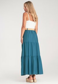 Długa, warstwowa spódnica wykonana z materiału w kolorze teal, z elastycznym pasem i beżowym paskiem. Stworzona z prostym białym topem na ramiączkach i sandałami na platformie.