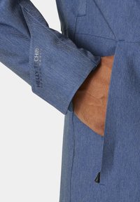 Manica di giacca in denim blu con logo Helly Tech®, con cuciture visibili, apertura della tasca e primo piano di un tessuto testurizzato.