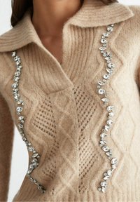 Beżowy sweter typu cardigan z dzianiny, z ribbanym kołnierzem, prezentujący wzór w romby i ozdobiony przezroczystymi kryształkami na przodzie.